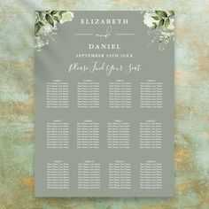 a wedding seating chart with white flowers and greenery on grey paper, sitting on a green background