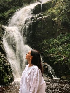 a woman is standing in front of a waterfall and looking up into the sky with her eyes closed