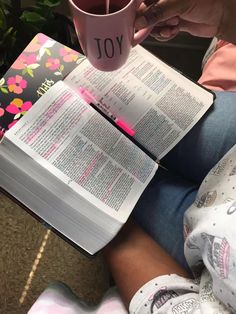 a person sitting on the floor reading a book and holding a coffee mug in their hand