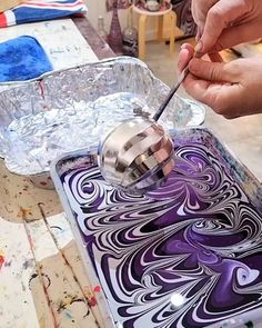 a person is painting with purple and white swirls on a sheet of paper while another hand holds a paintbrush