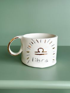 a white ceramic cup with a gold handle on a green surface, that reads libra