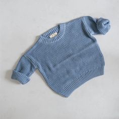 A soft, blue knit sweater with a round neckline and rolled-up sleeves is laid flat on a light gray surface. The sweater features a ribbed hem and cuffs, adding texture to its simple design. Knit Baby Sweater, Chunky Oversized Sweater, Oversized Knit Sweater, Knit Baby Sweaters, Baby Sweater, Oversize Knit, Oversized Knitted Sweaters