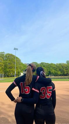 two female softball players in black uniforms standing on a baseball field with their arms around each other