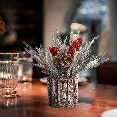 a wooden table topped with a vase filled with red flowers and pineconi branches