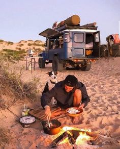 a man sitting in the sand next to a campfire with his dog looking on