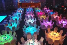 a room filled with lots of tables and chairs covered in white tablecloths, lit up by colorful lights