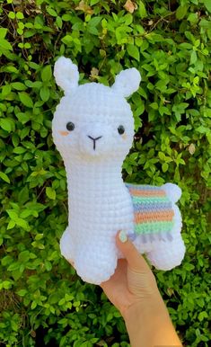 a hand holding up a small white llama toy in front of green bushes and shrubbery