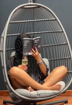 a woman sitting in a chair reading a book and holding a wine glass up to her face