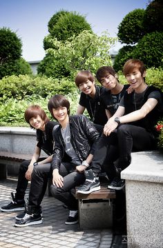 four boys are sitting on a bench in front of some bushes and trees, one boy is wearing a black t - shirt