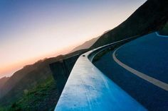 a curved road on the side of a mountain with mountains in the background at sunset