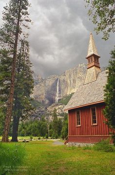 a small red church with a steeple in front of a mountain range and trees