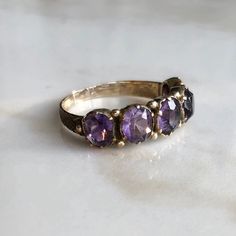 A late Georgian (circa.1830-1837) 15 carat gold Amethyst ring. This beautiful piece holds all five stones neatly in line, what a gorgeous collection of glowing purple stones. The shoulders show a nice bit of detailing too. CONDITION: Wear consistent with age and use. Centre stone previously replaced. Please see photos for more detail. SETTING (ACROSS): 30mm STONE DIAMETER: 5mm BAND WIDTH (SMALLEST): 2mm RING SIZE: UK: N 1/2 | US: 6 3/4 WEIGHT: 2.4 grams (SZZ) Heirloom Purple Ring With Rose Cut Diamonds, Victorian Amethyst Ring For Anniversary, Victorian Style Purple Amethyst Ring For Anniversary, Heirloom Multi-stone Amethyst Ring, Antique Oval Amethyst Multi-stone Ring, Victorian Amethyst Ring In Yellow Gold, Victorian Yellow Gold Amethyst Gemstone Ring, Classic Oval Multi-stone Amethyst Ring, Victorian Three Stone Oval Ring