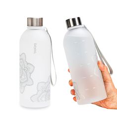 a hand holding a white water bottle next to a silver and black flask with an intricate design on it