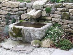 a stone fountain with moss growing on it in front of a rock wall and flowers