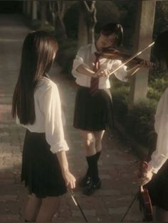 three girls in school uniforms are playing violin on the sidewalk while one girl is wearing a white shirt and black skirt