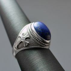 This Sterling Silver Fleur De Lis Cross Lapis Lazuli Oval Gemstone Ring is a stunning blend of elegance and historical charm. Featuring a dark blue lapis lazuli gemstone, known for its protective and intuitive properties, set in a finely crafted sterling silver band. The intricate Fleur De Lis Cross design adds a regal touch, making it perfect for both everyday wear and special occasions. Whether you're treating yourself or looking for a unique gift, this ring is a timeless addition to any jewelry collection. Metal: 925 Sterling Silver (stamped). Lapis Lazuli, with its deep blue hue and golden pyrite flecks, has been prized for centuries for its mesmerizing beauty and spiritual significance. In jewelry, Lapis Lazuli is believed to promote inner peace, clarity of mind, and self-expression. Classic Round Lapis Lazuli Jewelry, Classic Sapphire Oval Cabochon Jewelry, Elegant Blue Oval Cabochon Signet Ring, Classic Blue Oval Cabochon Signet Ring, Classic Blue Lapis Lazuli Rings, Classic Lapis Lazuli Jewelry Ring, Classic Rings With Natural Stones, Blue Lapis Lazuli Oval Jewelry, Classic Royal Blue Oval Jewelry