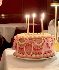 a pink birthday cake with lit candles on it sitting on a white table cloth covered table