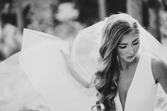 a woman wearing a wedding dress and veil looking down at her hair while standing in front of trees