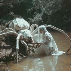 a woman in white dress standing next to a giant spider sitting on top of a body of water