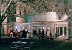 a group of people standing outside of a building at night with tables and chairs in the foreground