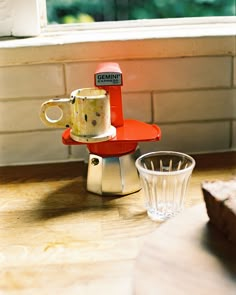 a red coffee maker sitting on top of a wooden table next to a glass cup