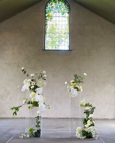 three clear vases with white flowers and greenery in front of a stained glass window