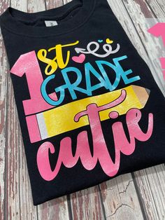 a black tshirt with the words 1st grade cutie written in pink and blue