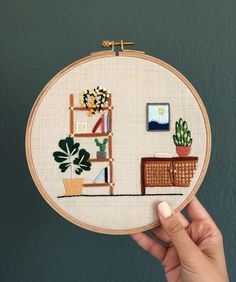 a hand holding up a cross stitch embroidery hoop with plants and bookshelves on it