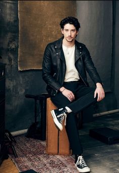 a man in black jacket and white shirt sitting on wooden stool with his legs crossed