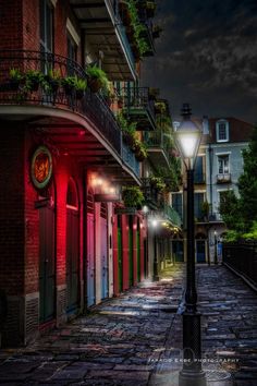 an empty street at night with red lights on the buildings and green plants hanging from balconies