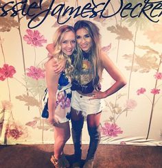 two beautiful young women standing next to each other in front of a wall with flowers