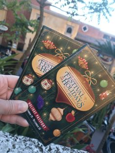 someone holding up two holiday cards in front of a building with lights and decorations on it