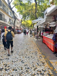 people walking down a cobblestone street with shops on both sides and trees in the background