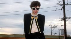 a woman wearing sunglasses and a black suit with yellow details on her neck, standing in front of power lines