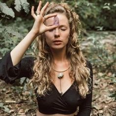 a woman with long hair holding her hand up to her face while standing in the woods