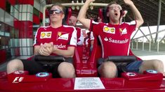 two men in red shirts sitting on a roller coaster at an amusement park with their hands up