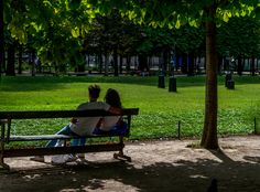 two people are sitting on a bench in the middle of a park, one is holding his arm around the other's back