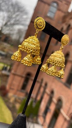 These earrings are traditional Indian jhumkas, characterized by their dome-shaped design. They are made of gold-toned metal and feature intricate detailing throughout. The top part of each earring is a circular stud with an ornate pattern, which is connected to the main jhumka portion by a small link. The main jhumka portion is highly detailed with layers of intricate patterns and decorative elements. Below the dome, there are smaller, dangling elements that add movement and additional embellish Small Gold Jhumka, South Indian Earrings, Temple Earrings, Gold Jhumka, Ornate Pattern, Gold Jhumka Earrings, Temple Jewelry, Earrings Indian, Traditional Earrings