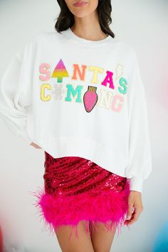 a woman wearing a santa's coming sweatshirt with pink feathers on the bottom and red sequin skirt