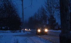 two cars driving down a snowy road at night with headlights on and snow covered trees in the background