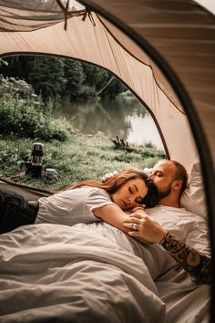 a man and woman laying in bed under a tent next to a lake with trees