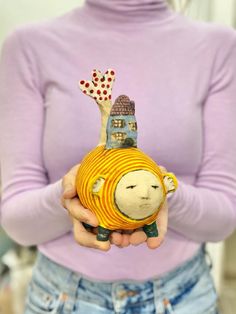 a woman holding a small yellow object with a house on it's head and a tree sticking out of the top