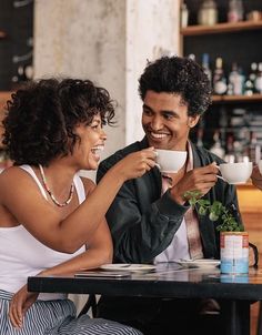 two people sitting at a table drinking coffee and talking to each other while holding cups in their hands