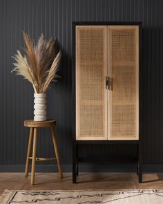 a tall wooden cabinet sitting next to a plant on top of a stool in front of a black wall