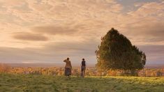 two people standing on top of a lush green field next to a tree in the distance