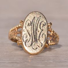 a gold ring with an ornate monogrammed design on the front and side, sitting on a wooden surface