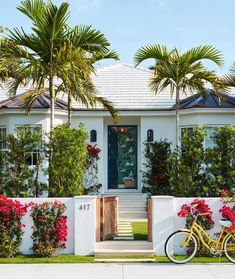 a yellow bike parked in front of a white house with red flowers and palm trees