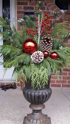 a vase filled with pine cones and greenery on top of a sidewalk next to a brick building