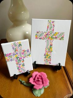 two cross paintings sitting on top of a wooden table next to a pink flower and a white vase