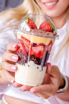 a woman holding up a smoothie with strawberries on top in a plastic cup